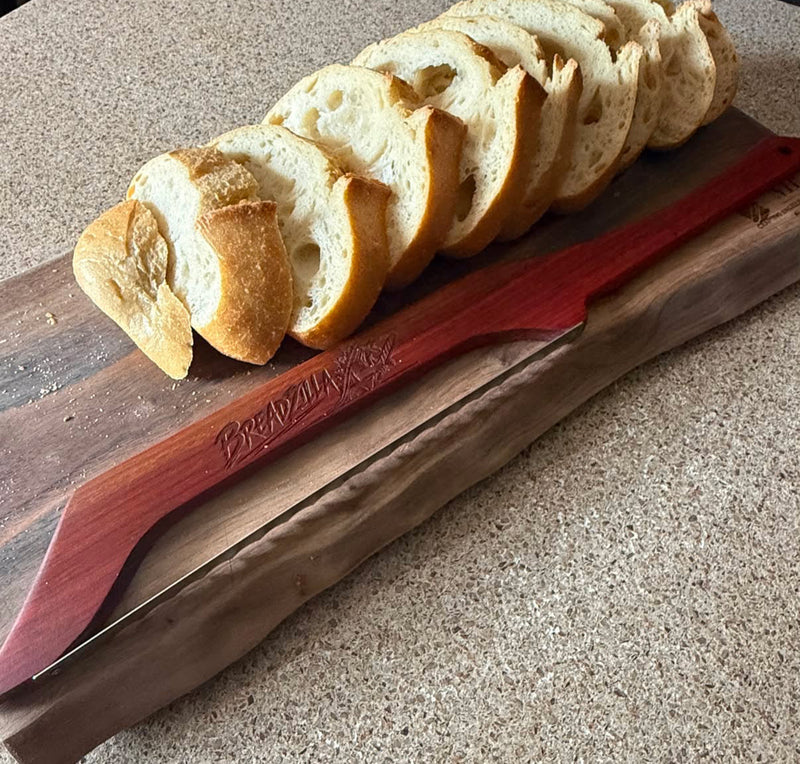 A wooden cutting board with a red handle knife, shown on a speckled countertop with sliced bread on top.