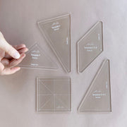 Five clear acrylic triangle and square templates labeled with template names are arranged on a light taupe background. A hand is holding one triangular template on the left.
